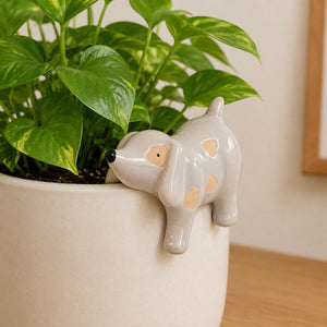 A small glossy ceramic dog planter in light gray with peach patches, shown growing in a white pot with green plant leaves against an indoor wall - Dog Pot Hanger Bisque -  6cmbackground.