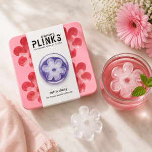 Drinks Plinks - Retro Daisy Pink Ice Cube Tray, wrapped in a white paper band labeled 'DRINKS PLINKS', shown on a light marble surface next to a glass of pink drink with a flower-shaped ice cube and pink flowers.