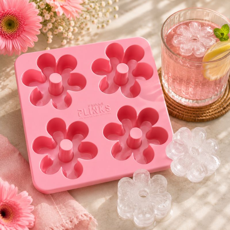 Drinks Plinks - Retro Daisy Pink Ice Cube Tray, shown on a light surface with pink flowers, a glass of pink drink, and clear flower-shaped ice cubes. The mold has the brand name PHILIPPE FLINKS printed on it.