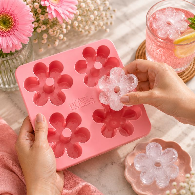 Drinks Plinks - Retro Daisy Pink Ice Cube Tray is shown on a marble surface, with two hands holding a clear flower-shaped ice cube over the mold. A pink drink in a glass and flowers are styled in the background.