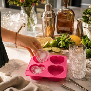 A hand is shown molding a Drinks Plinks -Big Heart Pink Ice Cube Tray on a marble countertop, with limes, a shaker, a bottle, and a glass nearby.