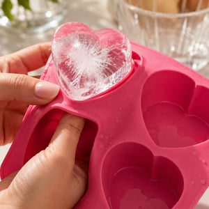 A close-up of hands holding a Drinks Plinks - Big Heart Pink Ice Cube Tray with one heart cavity filled with a clear ice heart and being lifted by fingers.