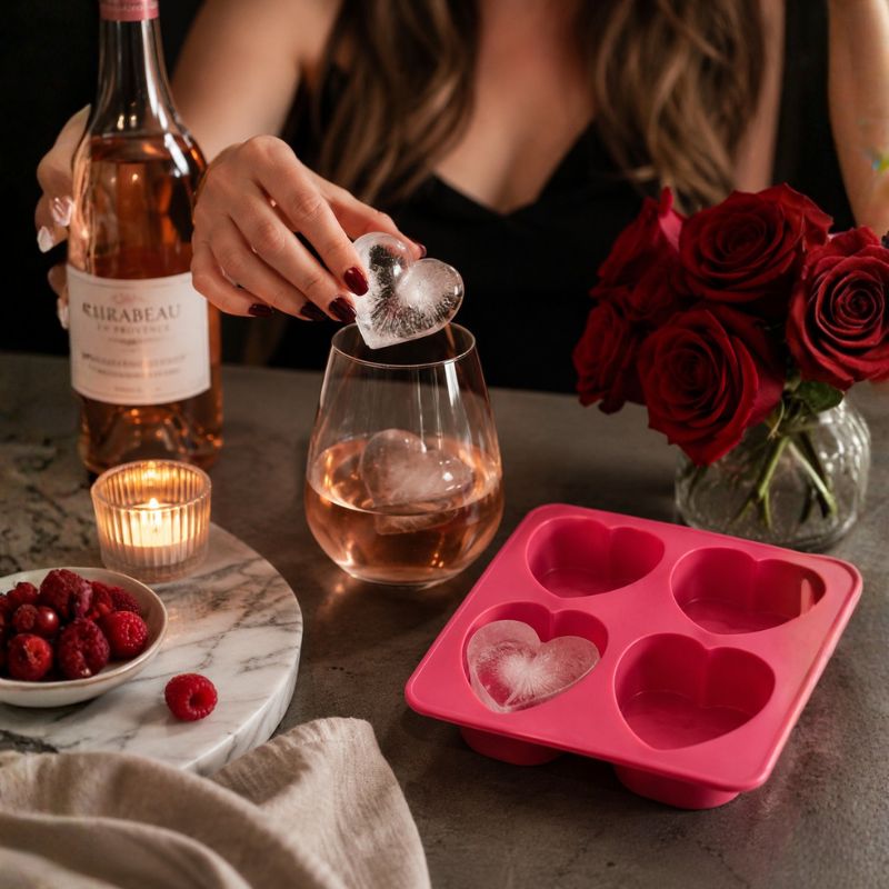 A Drinks Plinks -Big Heart Pink Ice Cube Tray sits on a table next to a bottle of Rosebeau rosé wine, a glass of rosé with ice, red roses, and a person's hands pouring a heart-shaped ice cube into a glass.
