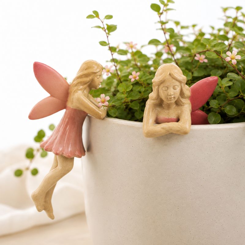 Two small fairy garden figurines with pink wings are placed on a white planter filled with green leafy plants. One fairy is leaning against the pot holding a small pink flower, and the other is sitting inside the pot with arms crossed.-