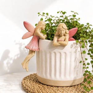A white ribbed ceramic planter with two decorative fairy figurines perched on the rim, filled with green trailing foliage, set on a woven round mat against a white background.