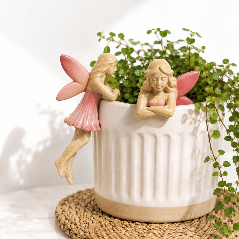 A white ribbed ceramic planter with two decorative fairy figurines perched on the rim, filled with green trailing foliage, set on a woven round mat against a white background.