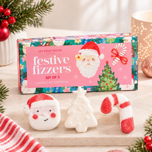 Annabel Trends Festive Fizzers set of 3 bath bombs displayed on a light stone surface with holiday decorations. The colorful box features Santa, a Christmas tree, and a candy cane, while the three bath bombs in front are shaped like Santa's face, a Christmas tree, and a candy cane.