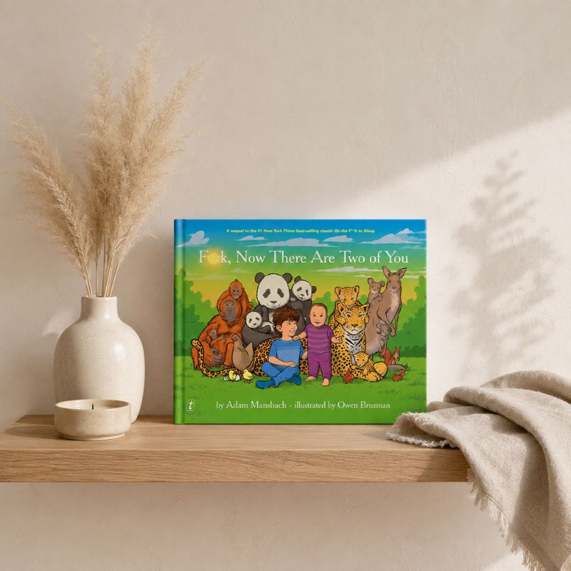 A children's board book titled "Fork, Now There Are Two of You" sits on a wooden shelf in a styled interior setting. The cover shows a colorful illustration of two children surrounded by animals, including a panda, tiger, lion, elephant, and goat.