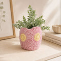 Pink ceramic vase with floral patterns containing a plant on a surface with books and a framed picture in the background.