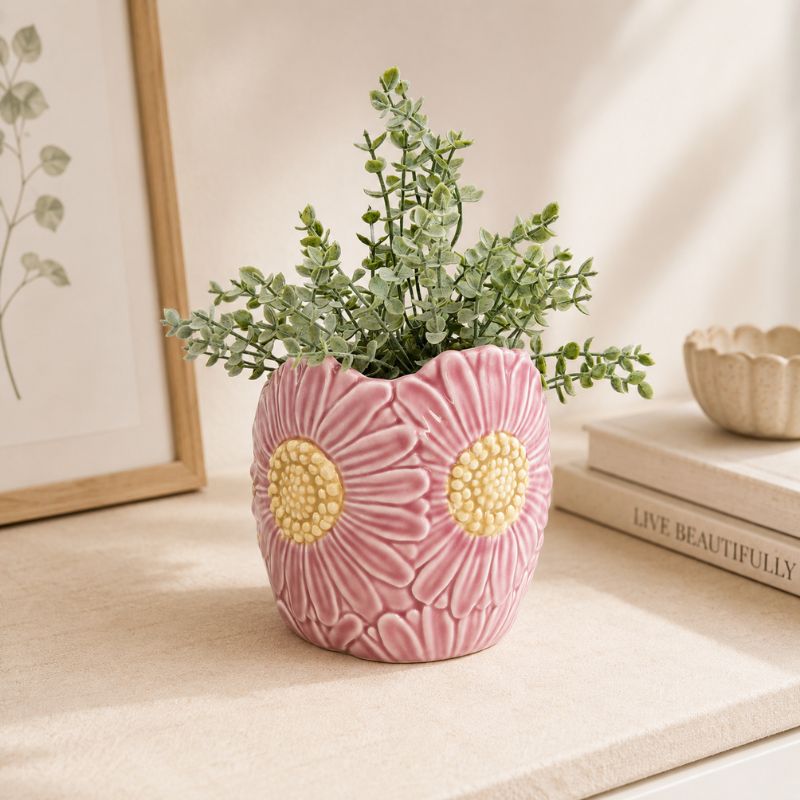 Pink ceramic vase with floral patterns containing a plant on a surface with books and a framed picture in the background.