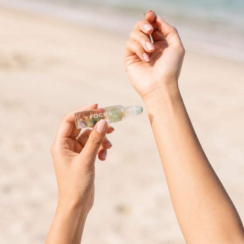 Two hands holding a small transparent container with 'FOCUS rollerball bottle' against a blurred beach background.