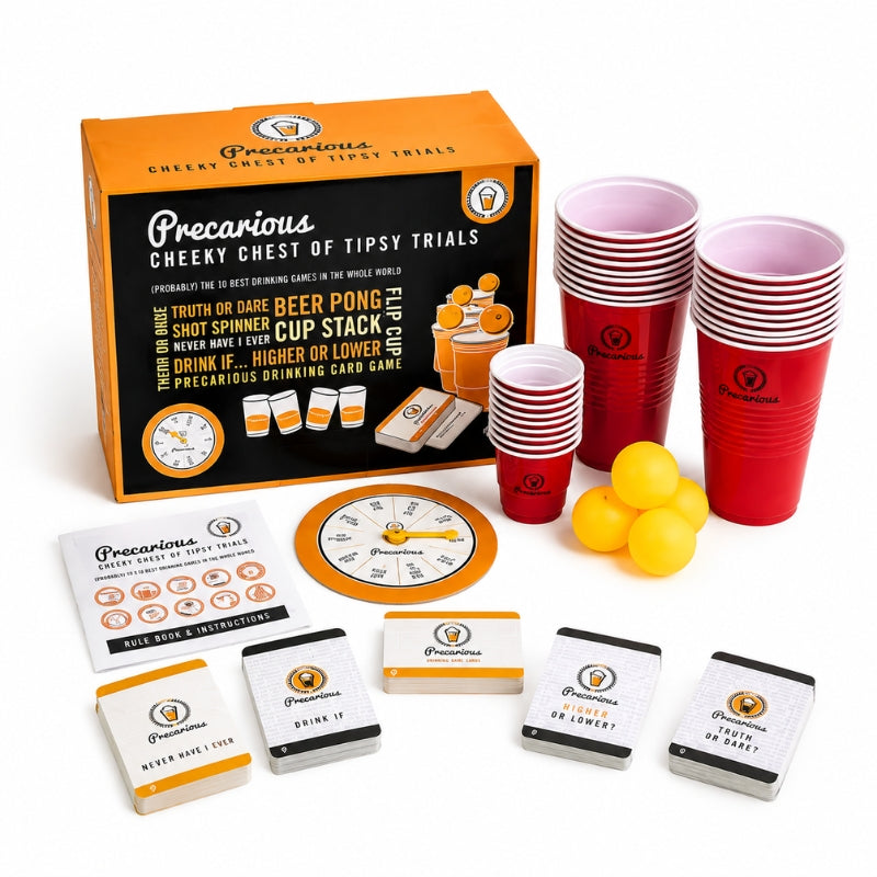 Precarious Cheeky Chest of Tipsy Trials drinking game set with orange and black retail box, red plastic cups, yellow balls, cards, spinner, and other game pieces on a white background.
