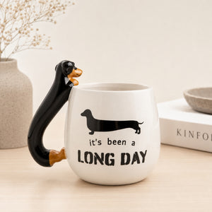 A Furever Pets Dachshund 3D Handle Mug with a black dachshund-shaped handle and printed black text reading 'It's been a LONG DAY' on the front, styled on a light wood surface with a vase and books in the background.