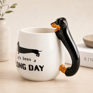 Furever Pets Dachshund 3D Handle Mug with a dachshund-shaped handle in black and tan, printed with a black dachshund silhouette and the text 'It's been a LONG DAY' on a light wood tabletop.