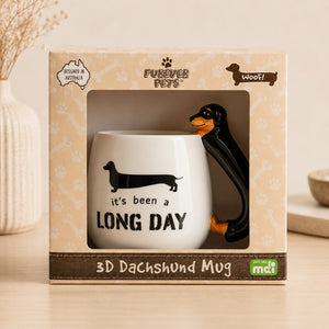 A 3D Furever Pets Dachshund 3D Handle Mug by Forever Pets, displayed inside its original packaging. The mug features a white body with a black dachshund silhouette and the text 'It's been a LONG DAY,' while the handle is shaped like a black and tan dachshund.