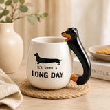 A Furever Pets Dachshund 3D Handle Mug with a black dachshund-shaped handle and the text 'It's been a LONG DAY' printed on the front, styled on a woven coaster in a warm home setting.