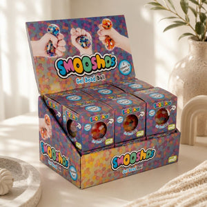 A display box of SmooShos Gel Bead Ball candy, shown in a bright, sunlit room with a neutral background and a decorative plant in a vase.