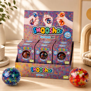 A display box of Gel Bead Multi Smooshos Balls with three individual packs in front, styled on a wooden surface with two colorful gel balls in the foreground and a plant in the background.
