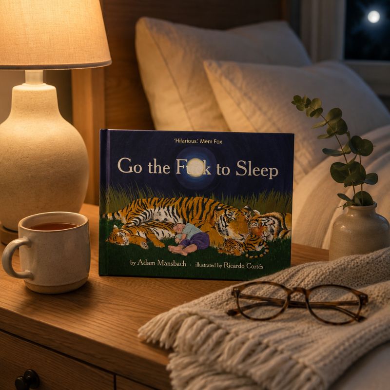 A children's board book titled "Go the F*ck to Sleep" by Adam Mambach and Ricardo Cortés sits on a wooden bedside table in a cozy bedroom setting. The book cover features an illustration of a tiger sleeping on a bed next to a person.