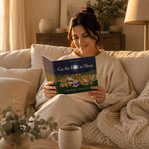 A woman sits on a beige sofa in a warmly lit living room, reading a book titled "Go the F*ck to Sleep" with a tiger illustration on the cover.