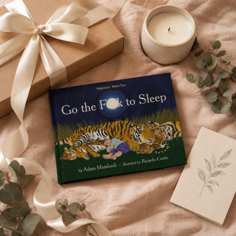 A children's book titled "Go the Fox to Sleep" by Hilarious Mom Fox is styled in a cozy flat lay on a soft beige fabric, surrounded by a wrapped gift with a cream ribbon, a lit candle, eucalyptus leaves, and a small card.