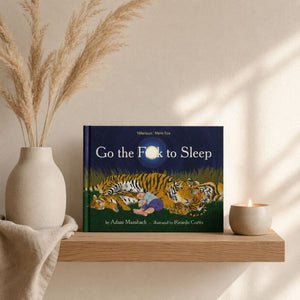 A children's book titled "Go the F*ck to Sleep" by Adam Munsch and Ricardo Cortés sits on a wooden shelf in a styled interior scene. The book cover shows a dark blue night sky with a tiger and a person under a full moon.