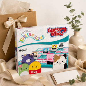 Guess Who? Squishmallows game box displayed on a styled tabletop with a kraft gift box, cream ribbon, candle, eucalyptus sprigs, and a blank card. The box shows colorful cartoon characters and the Guess Who? branding.