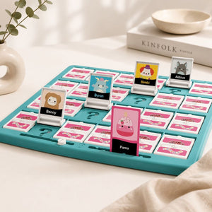A Guess Who - Squishmallows Edition Board Game on a light tabletop with decorative props including a vase with greenery, a book labeled KINFOLK, and a small bowl. The board is teal with pink and white cards, and several character cards are standing up.