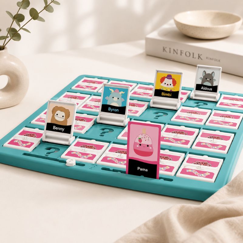 A Guess Who - Squishmallows Edition Board Game on a light tabletop with decorative props including a vase with greenery, a book labeled KINFOLK, and a small bowl. The board is teal with pink and white cards, and several character cards are standing up.
