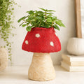 A HUSK Toadstool Planter – Red & White 19cm made from red and beige yarn, with a green potted plant inside, photographed on a aesthetic background.