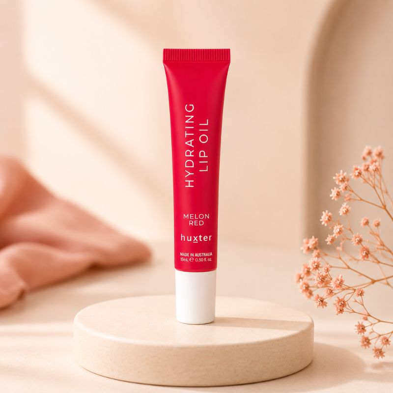 Huxter Melon Red Hydrating Lip Oil tube on cream pedestal with coral blush styling