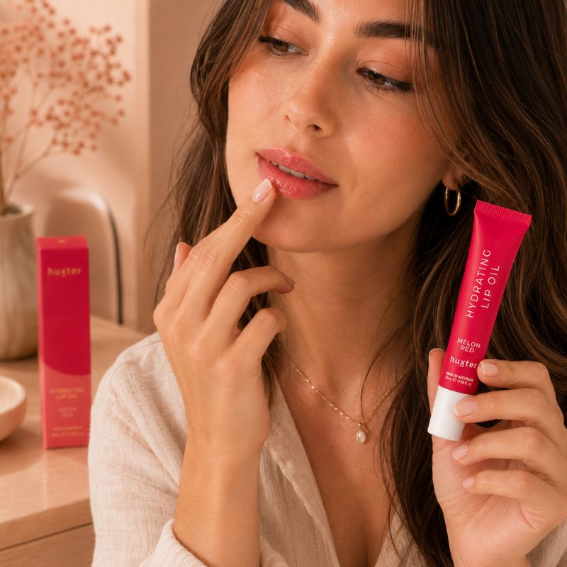 Woman applying Huxter Melon Red Hydrating Lip Oil with fingertip in lifestyle setting