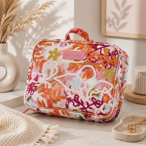 A Large hanging toiletry bag in Ivy floral print with pink and orange details with a colorful floral and abstract botanical print, featuring a pink top handle and a pink brand patch on the front, styled on a light stone surface with dried flowers and home decor props.
