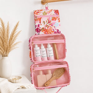 A Large hanging toiletry bag in Ivy floral print with pink and orange detailshangs from a wooden rod on a white wall. The open bag has clear pockets containing three white bottles, a wooden stick, and two pink bottles, with a wooden brush resting inside.