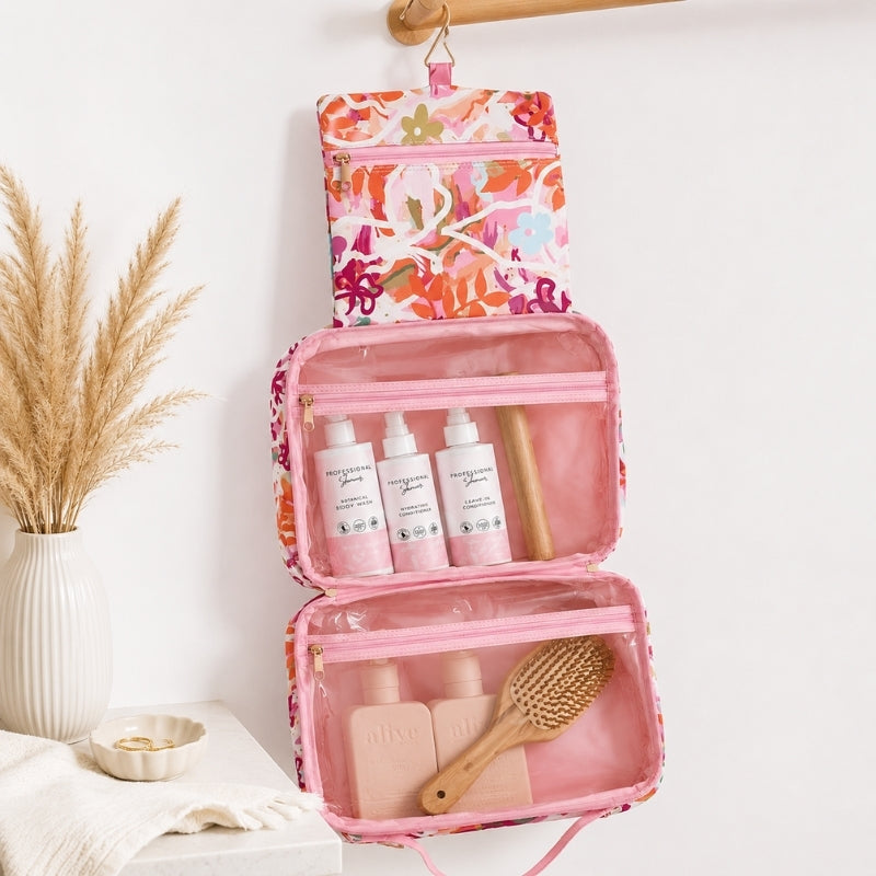 A Large hanging toiletry bag in Ivy floral print with pink and orange detailshangs from a wooden rod on a white wall. The open bag has clear pockets containing three white bottles, a wooden stick, and two pink bottles, with a wooden brush resting inside.