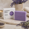 Lavender soap bar with packaging on a wooden surface with lavender flowers.