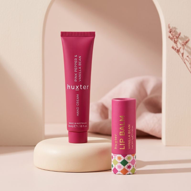 Huxter Pink Pepper and Vanilla Bean hand cream with Vanilla Bean lip balm on a warm blush studio pedestal