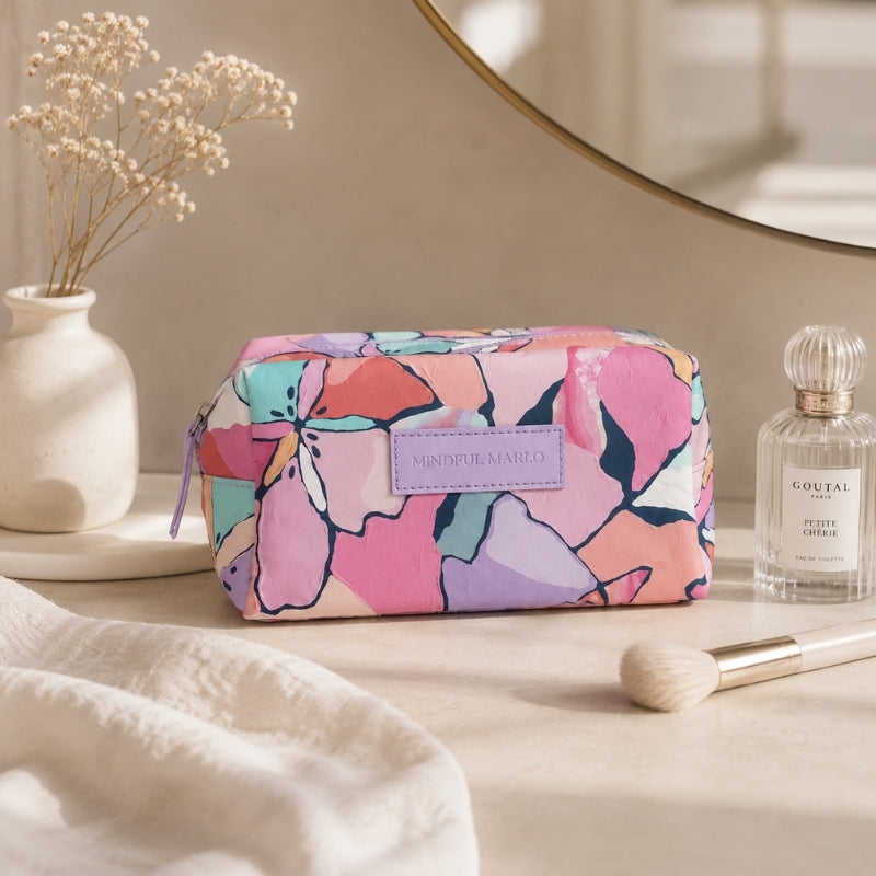 A Makeup Cosmetic Bag – Willow with a colorful floral pattern in pink, purple, teal, and coral, styled on a beige tabletop next to a perfume bottle, a makeup brush, and dried flowers in a vase.