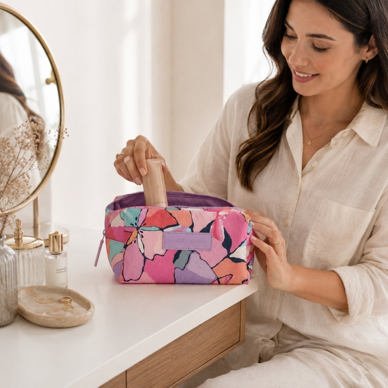 A woman in a white linen shirt sits at a vanity table, placing a makeup sponge into a Makeup Cosmetic Bag – Willow. The pouch features a vibrant pink, purple, and orange abstract floral pattern with a purple zipper and a lavender brand tab.