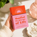 A hand holds a pink and orange card deck box with the text 'Manifest the Shit Out of Life' in a bright, styled setting with a plant, a copper-toned vase, and jewelry in the background.