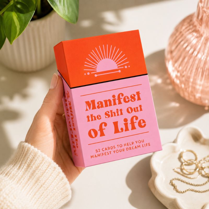A hand holds a pink and orange card deck box with the text 'Manifest the Shit Out of Life' in a bright, styled setting with a plant, a copper-toned vase, and jewelry in the background.