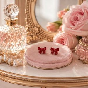 Mini Bow Stud Earrings on a pink cushion with a perfume bottle and flowers in the background.