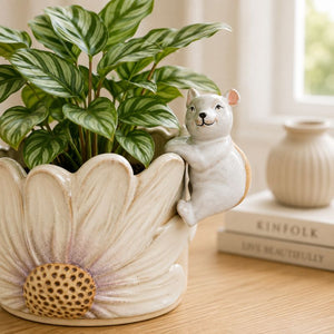A ceramic planter shaped like a large cream-colored flower with a small white cat figurine perched on the side, holding a green leafy plant. The planter sits on a wooden table in a softly lit room, with books and another vase blurred in the background.-Mouse Pot Hanger White 9cm