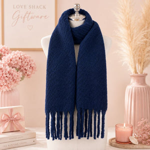 A Navy Blue Chunky Knit Winter Scarf with Fringe displayed on a white mannequin torso, styled in a soft pink gift-themed setting with pink flowers, a vase, a candle, and a gift box.