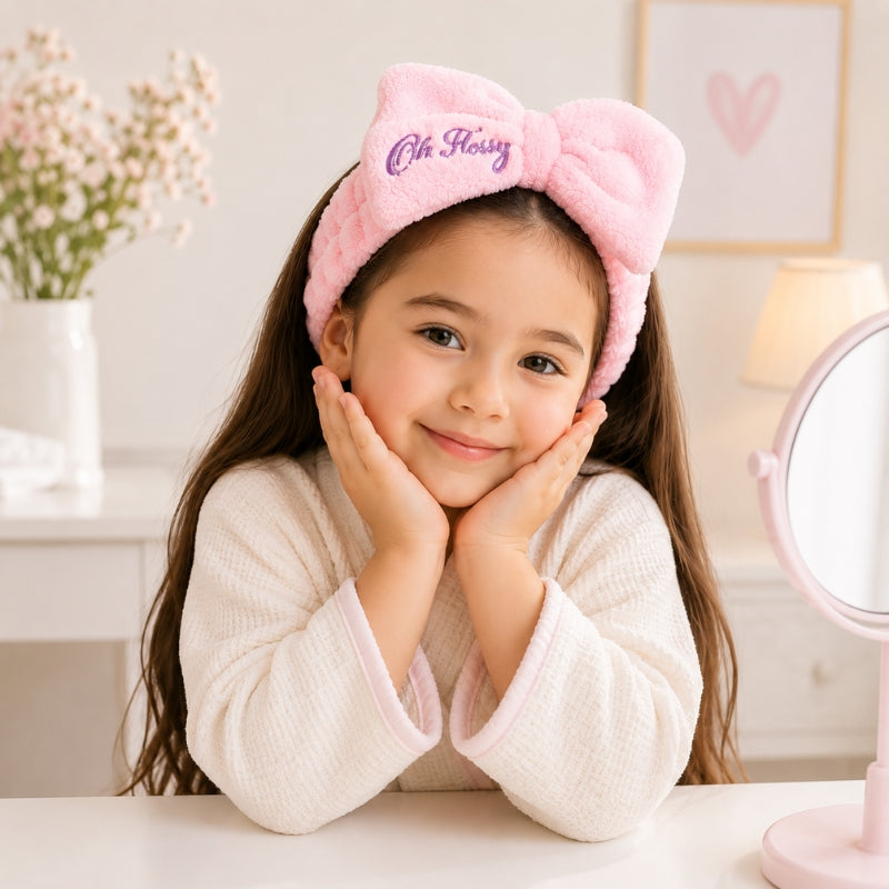 A young girl with long brown hair smiles while wearing a pink plush bow headband with 'Oh Flossy' embroidered in purple on it, sitting at a white table in a bright, soft room.