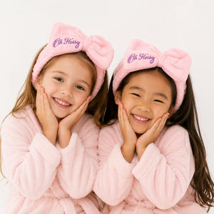Two young girls smiling and posing together, wearing matching pink plush bow headbands with the name 'Oh Flossy' embroidered in purple on the front, paired with light pink bathrobes against a clean white background.