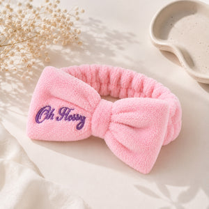 A pink plush bow headband with purple embroidered text reading 'Oh Flossy' on the front, styled on a light cream surface with dried flowers and a ceramic dish in the background.