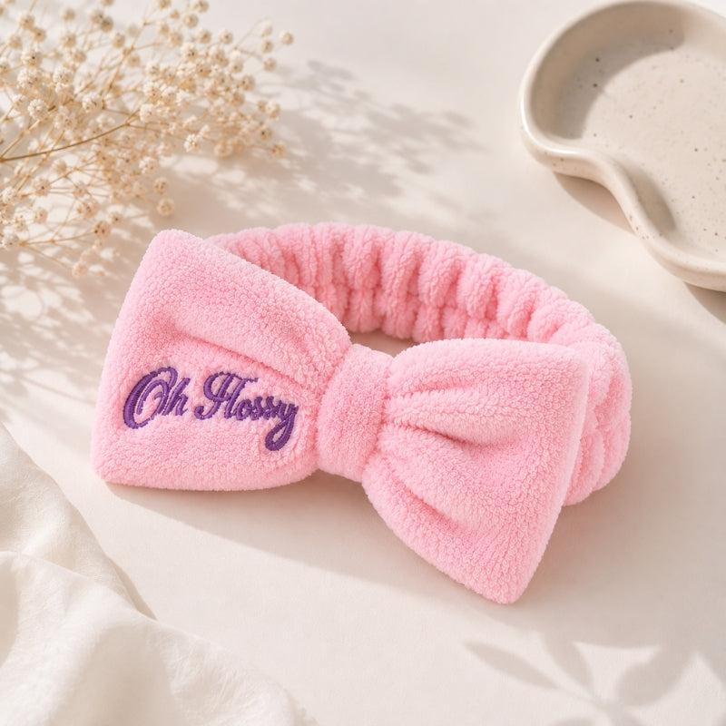 A pink plush bow headband with purple embroidered text reading 'Oh Flossy' on the front, styled on a light cream surface with dried flowers and a ceramic dish in the background.