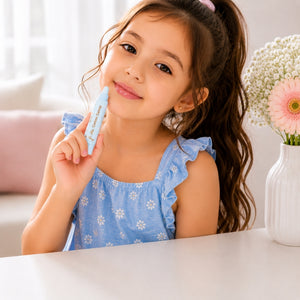 A young girl with long brown hair in a ponytail smiles while holding a Young girl using Oh Flossy Cotton Candy kids natural lip gloss. She is wearing a light blue ruffled top with white daisy patterns.