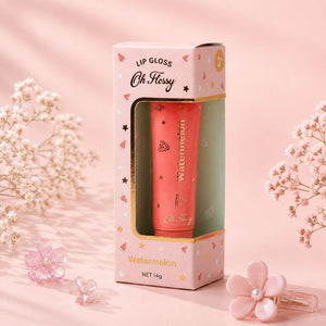 A pink lip gloss tube labeled 'Oh Hissy' and 'Watermelon' displayed inside its matching pink packaging box, styled with pink flower props on a soft pink background.